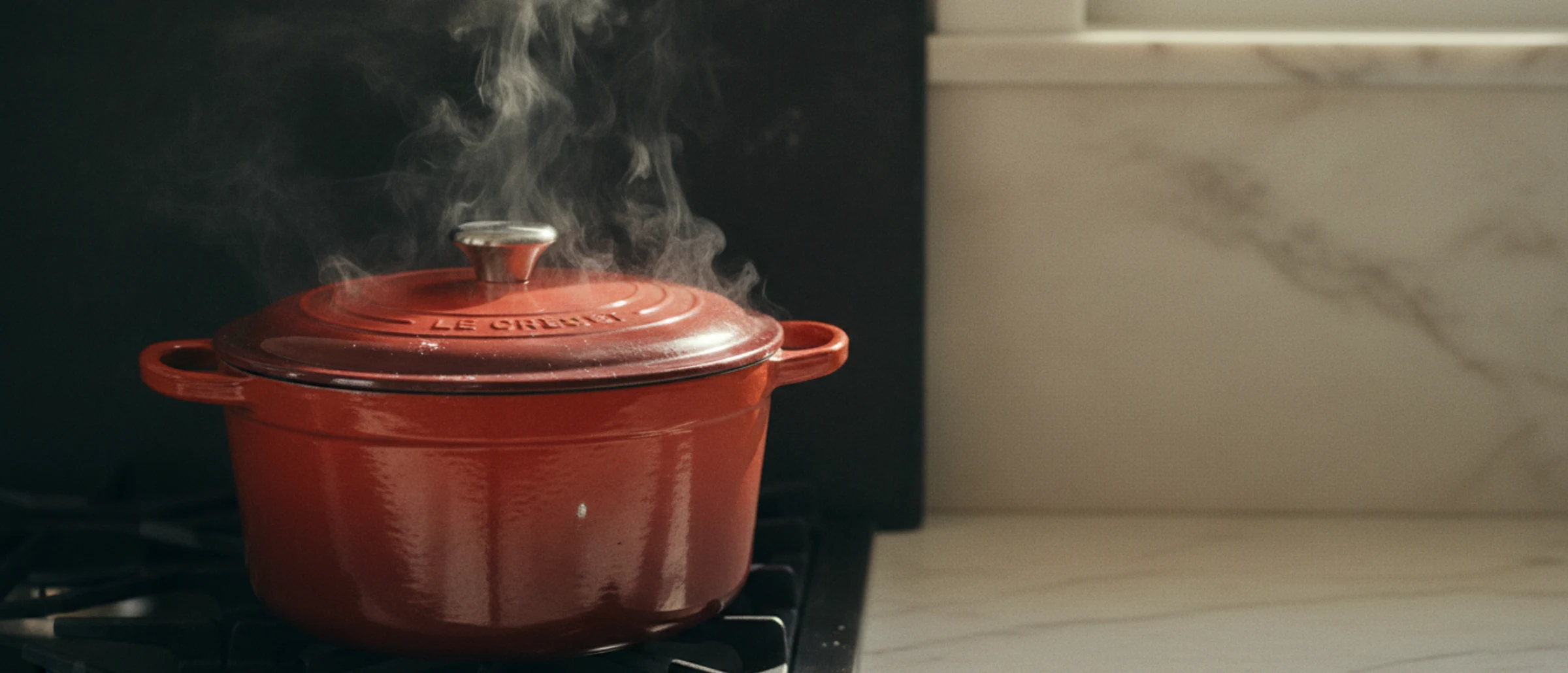 Dutch oven Lodge rojo y sartén All-Clad de acero inoxidable sobre piedra negra, con fondo oscuro liso a la derecha