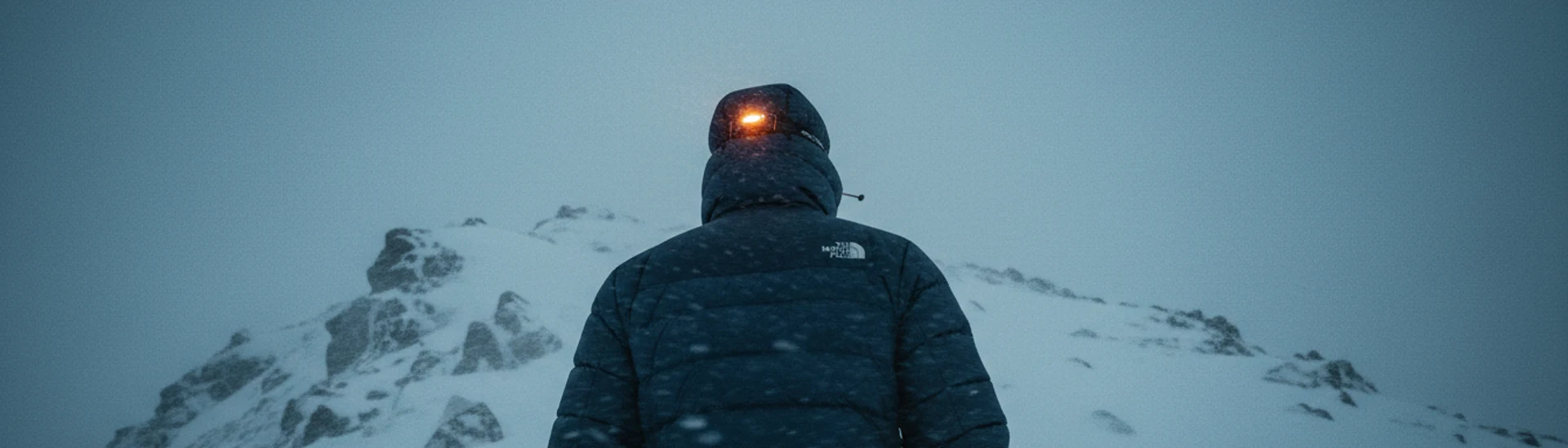 Detalle de una parka técnica con capucha de piel sintética, gotas de nieve sobre la tela y una mano ajustando el cierre, con montaña nevada de fondo.